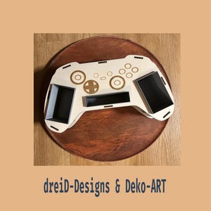 Könnte beinhalten: Ein Gamecontroller-Organizer aus hellem Holz mit lasergravierten Tastendetails, einem zentralen rechteckigen Schlitz und zwei Seitenfächern mit dunklem Innenraum. Der Text "dreiD-Designs & Deko-ART" ist unter dem Objekt aufgedruckt.