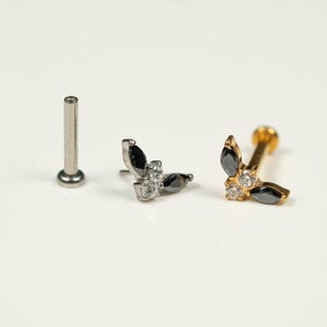 Könnte beinhalten: Drei Schmuckstücke werden gezeigt. Zwei Ohrringe mit Blattdesign, einer silberfarben und einer goldfarben, jeweils mit schwarzen und klaren Edelsteinen. Ein silberner Stecker ist ebenfalls enthalten.