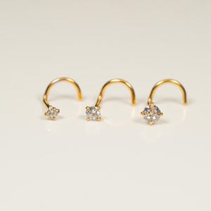 May include: Three gold nose rings with a small clear gemstone on each. The rings are curved and have a small loop at the end.