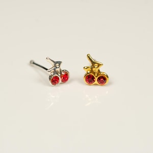 May include: Two cherry-shaped nose studs. One is silver-colored with red gems, and the other is gold-colored with red gems. The studs are small and delicate, with a polished finish. The background is a plain white surface.