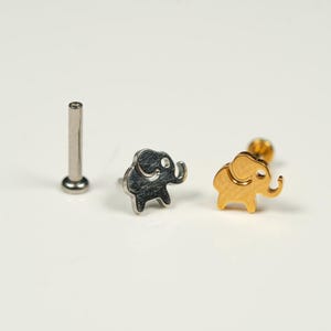 Pode incluir: Três anéis de nariz de elefante em prata e ouro. Os anéis são pequenos e delicados, com um design simples. O anel de prata está à esquerda, o anel de ouro está à direita e o poste de prata liso está no centro.