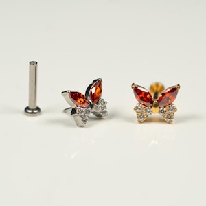 May include: Two butterfly-shaped body jewelry pieces with red and clear rhinestones. One piece is gold-toned and the other is silver-toned. A silver-toned post is also shown.