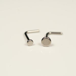 May include: Two silver nose rings with a round, flat top. The nose ring on the left has a smaller top, while the nose ring on the right has a larger top.