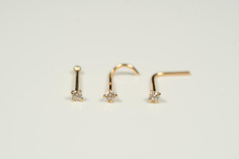 Puede incluir: Tres anillos de nariz de oro con una peque&ntilde;a piedra preciosa transparente en cada uno. Los anillos est&aacute;n en una fila sobre una superficie blanca.
