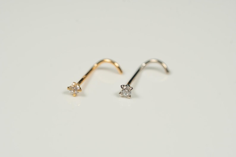 Puede incluir: Dos anillos de nariz con un peque&ntilde;o diamante engastado en garras. El anillo de nariz de la izquierda es de oro y el anillo de nariz de la derecha es de plata.