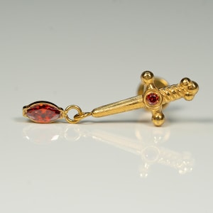 Puede incluir: Pendiente de espada de oro con un colgante de piedra roja. La espada tiene un dise&ntilde;o retorcido y una piedra roja en la parte superior.
