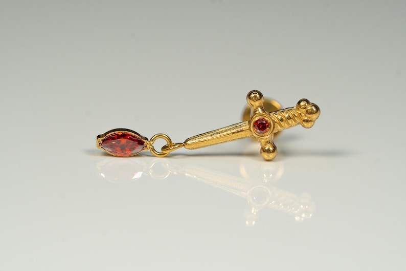 Puede incluir: Pendiente de espada de oro con un colgante de piedra roja. La espada tiene un dise&ntilde;o retorcido y una piedra roja en la parte superior.