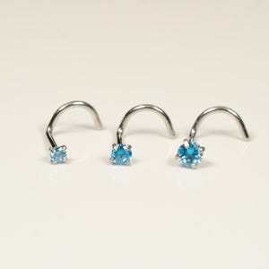 Peut inclure: Trois anneaux de nez argentés avec une pierre précieuse bleue sur chacun. Les anneaux sont incurvés et ont une petite boucle à l'extrémité.