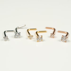 May include: An assortment of nose studs in silver, gold, and rose gold. Each stud features a small, clear, round gemstone. The L-shaped posts are visible, and the jewelry is arranged on a white surface.