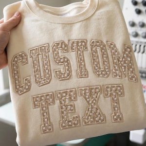 May include: A cream-colored crewneck sweatshirt featuring the words CUSTOM TEXT in large, block-style letters. The letters are appliqued with a tan fabric that has a white polka dot pattern and are outlined with a visible stitched border.