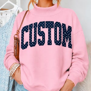 May include: A light pink long-sleeve sweatshirt with a mock neck collar. The front features the word CUSTOM in large, dark navy blue block letters with a white polka dot pattern. The sweatshirt has long sleeves and a relaxed fit design.