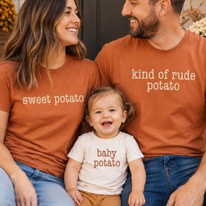 May include: A set of three t-shirts in rust orange and cream. The rust orange shirts feature the text "sweet potato" and "kind of rude potato" in white. The cream shirt features the text "baby potato" in rust orange lettering on the front.