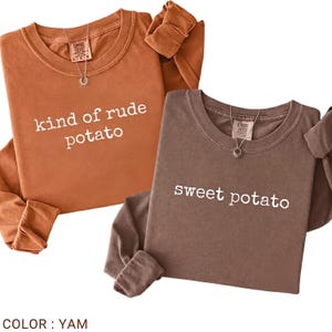 以下が含まれることがあります： ヤムと茶色の2色の長袖シャツ。ヤム色のシャツには「kind of rude potato」の文字、茶色のシャツには「sweet potato」の文字がプリントされています。どちらのシャツもクルーネックで、小さなペンダントネックレスが付いています。