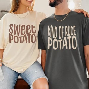 Op de afbeelding: Twee T-shirts, één crèmekleurig en één antracietgrijs, elk met een aardappelthema. Het crèmekleurige shirt zegt "Sweet Potato" in bruine tekst, terwijl het grijze shirt "Kind of Rude Potato" in witte tekst zegt. Beide zijn korte mouwen.