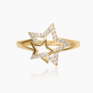 May include: A gold-colored ring featuring a star design. The star is outlined with small, clear gemstones. The ring has a double-star design, with one star inside the other. The band is a simple, smooth gold color.