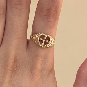 Peut inclure: Bague dorée avec un motif en forme de cœur et une croix au centre. La bague a une surface texturée et est portée au doigt. Il s'agit d'un bijou.
