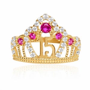 Pode incluir: Um anel dourado em forma de tiara com o número 15 ao centro. A banda é decorada com pedras brancas transparentes e cinco pedras redondas cor-de-rosa dispostas num padrão de coroa, com uma textura de contas ao longo da borda inferior.