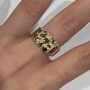 Peut inclure: Une bague dorée avec un motif texturé en forme de pépite. La bague est large et de forme rectangulaire, avec une surface détaillée qui capte la lumière. La bague est portée au doigt.