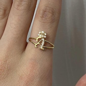 May include: A gold ring featuring a cursive letter A as the centerpiece. The letter A is embellished with small, clear, sparkling stones along its surface. Above the letter A sits a small, star-shaped cluster of clear stones, adding detail.