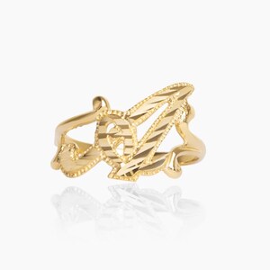 Puede incluir: Un anillo dorado con la letra "A" en una fuente cursiva. La letra tiene un diseño texturizado y cortado, mientras que la banda tiene un acabado liso. El anillo se muestra sobre un fondo blanco, resaltando su diseño.