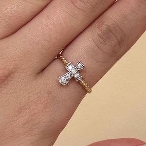 Peut inclure: Bague dorée ornée d'un pendentif en forme de croix incrusté de petites pierres scintillantes. L'anneau de la bague présente un motif torsadé. La croix est centrée sur le doigt.