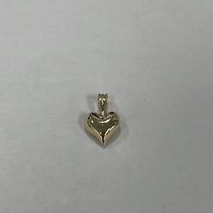 Peut inclure: Pendentif en forme de cœur doré, avec une petite boucle pour une chaîne. Le cœur est lisse et arrondi, avec une finition polie. Le pendentif est probablement en or ou plaqué or.