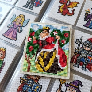 Puede incluir: Una colección de pequeños parches cuadrados de punto de cruz con personajes de fantasía en estilo pixel-art, incluyendo un rey, una reina, un caballero, un dragón y un mago. Cada parche se muestra en una funda de plástico transparente.