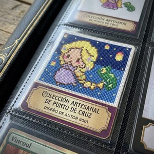 Puede incluir: Una tarjeta de patrón de punto de cruz con un personaje pixelado de cabello rubio largo y un camaleón verde sobre un cielo nocturno azul estrellado. La tarjeta incluye el texto Colección Artesanal de Punto de Cruz Diseño de Autor #001.