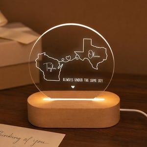 May include: A circular acrylic night light with a wooden base, featuring engraved outlines of Wisconsin and Texas connected by a line with a heart. The text reads Taylor, Olivia, and ALWAYS UNDER THE SAME SKY with a small heart below the text.