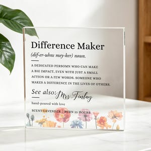 Könnte beinhalten: Eine quadratische Acrylplatte mit dem schwarz aufgedruckten Text Difference Maker, einer Definition und dem Namen Mrs. Finlay. Der untere Rand ist mit einer bunten Blumenillustration verziert, inklusive Scent: Lavender und Burn 25 Hours.