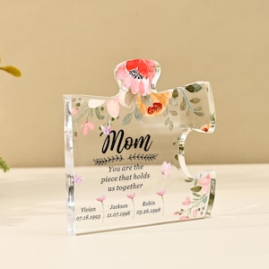 Puede incluir: Una pieza de rompecabezas de acrílico transparente con la palabra "Mom" y detalles florales. El texto "You are the piece that holds us together" está debajo de la palabra "Mom". Nombres y fechas están impresos en la parte inferior.