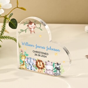 Pode incluir: Uma lembrança de acrílico transparente em forma de coração com um design de animais da selva. O texto "William James Johnson CHRISTENED 26.06.2024" está impresso em azul. Apresenta um macaco, uma girafa, um elefante, um leão, um hipopótamo e uma zebra.