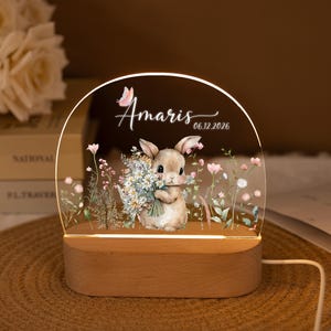 Może przedstawiać: Dekoracyjna lampka nocna z królikiem trzymającym kwiaty, z imieniem "Amaris" i datą "06.12.2026" wydrukowanymi na przezroczystym akrylu. Stoi na drewnianej podstawie i ma kwiatowe akcenty.