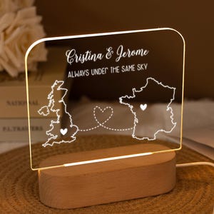 May include: A personalised night light with a wooden base. The clear acrylic panel displays the names "Cristina & Jerome" and the phrase "ALWAYS UNDER THE SAME SKY." Outlines of the UK and France are connected by a heart-shaped design.
