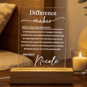 May include: A rectangular clear acrylic plaque set in a light wood base, featuring white engraved text that defines the word Difference maker as a special person who changes lives daily, followed by the name Nicole at the bottom.