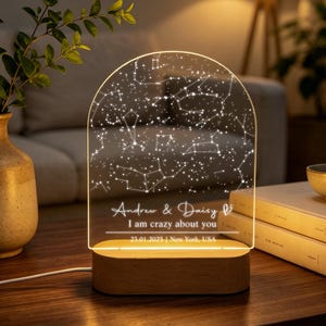Puede incluir: Una luz nocturna de acrílico arqueada con un diseño de mapa estelar blanco. La base es de madera clara. El texto dice: Andrew & Daisy, I am crazy about you, 23.01.2025 | New York, USA. La lámpara emite un brillo cálido sobre las constelaciones.