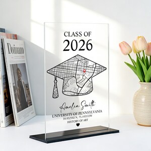 May include: Clear acrylic graduation plaque with a black base. The plaque features the text "CLASS OF 2026" above a graduation cap design with a map. Below the cap is the name "Amelia Smith", university details, and a heart symbol.