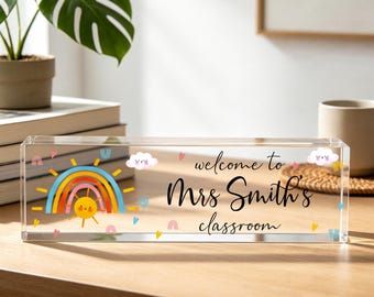 May include: A clear rectangular acrylic desk sign featuring a colorful rainbow and sun graphic on the left side, with the text "welcome to Mrs Smith's classroom" printed in black script, accented by small pink hearts and smiling clouds.