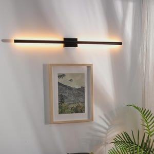 May include: A modern, black linear wall light fixture with a warm glow is mounted above a framed landscape painting. Below, a black leather sofa is adorned with decorative pillows featuring colorful geometric patterns and stripes. A potted plant adds a touch of greenery.