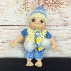 May include: A handmade doll in a blue and yellow crocheted outfit, including a hat, jacket, and shorts. The doll has a painted face with large eyes and a neutral expression. The outfit is decorated with pom-poms and buttons. The doll is standing on a black surface.