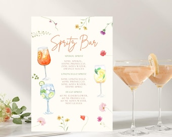 May include: A cream-colored Spritz Bar sign with watercolor illustrations of cocktails and floral accents. The sign lists recipes for Aperol, Limoncello, and Hugo Spritz cocktails. Two stemmed glasses with pink cocktails and lemon garnishes are in the foreground.