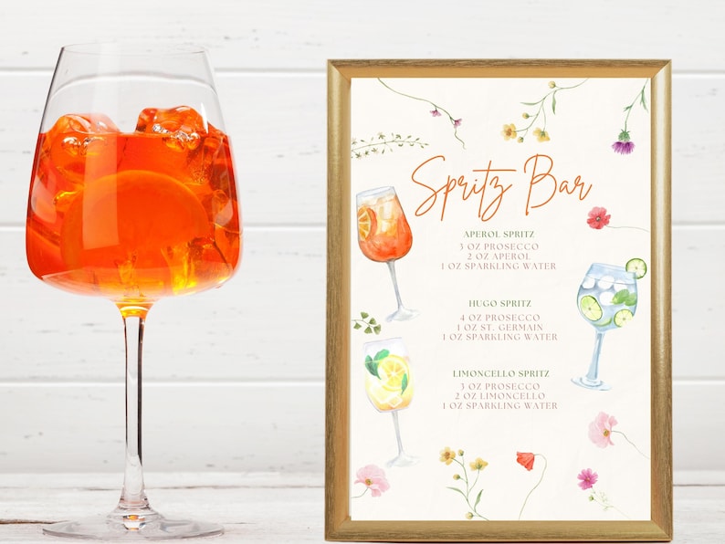 Puede incluir: Una copa de vino alta llena de un c&oacute;ctel naranja y cubitos de hielo, junto a un letrero enmarcado. El letrero dice "Spritz Bar" y muestra ilustraciones de c&oacute;cteles spritz con sus recetas.