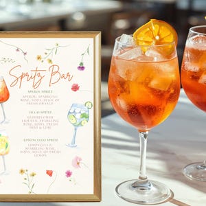Puede incluir: Un letrero con texto "Spritz Bar" e ilustraciones de c&oacute;cteles, junto a dos copas de c&oacute;cteles spritz de color naranja. El letrero detalla las recetas de spritz Aperol, Hugo y Limoncello. Los c&oacute;cteles tienen hielo y rodajas de naranja.