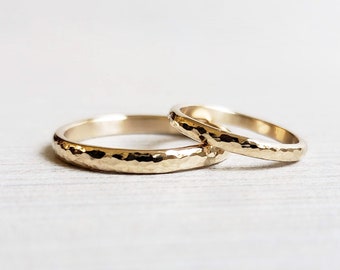 May include: Two gold hammered wedding bands, one slightly larger than the other, are stacked on a white surface.