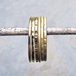 May include: A set of gold-coloured rings, with different textures, are arranged on a thin, light-brown twig. The rings have a hammered or textured finish, reflecting light. The background is a soft, blurred mix of blue and white.