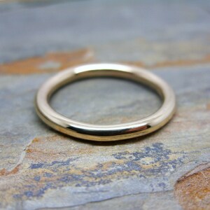 May include: A simple gold band ring, with a smooth, rounded surface. The ring is shown on a textured, brown surface.