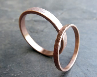 Peut inclure: Deux bagues de couleur cuivre entrelacées, présentant une texture martelée. Les bagues sont fines et ont une teinte brun-rougeâtre chaude. Le fond est gris clair, offrant un contraste aux bagues.