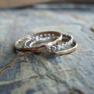 Könnte beinhalten: Drei goldene Ringe, die übereinander gestapelt sind. Der obere Ring hat eine strukturierte, gehämmerte Oberfläche. Der mittlere Ring hat eine Reihe kleiner silberner Perlen. Der untere Ring ist schlicht golden.