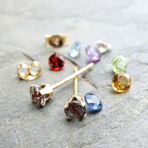May include: A pair of gold stud earrings featuring a dark green and blue gemstone. The earrings are surrounded by a selection of colourful gemstones, including red, blue, purple, green, and brown.