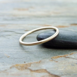 Op de afbeelding: Een dunne, goudkleurige ring ligt op een donkergrijze steen tegen een grijze, getextureerde ondergrond. De ring heeft een eenvoudig, minimalistisch ontwerp, met een gladde, afgeronde band. De ring lijkt van metaal te zijn en wordt als sieraad gedragen.